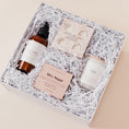 Load image into Gallery viewer, Lavender and Pine Self Care Sunday Gift Box angle view featuring Roote oat and honey body lotion, Thulisa Naturals shower steamer discovery set, NCLA Beauty Hey Sugar coconut vanilla body scrub, and Roote Amber Noir candle in white luxe two piece gift box