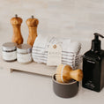 Load image into Gallery viewer, Lavender and Pine Well-Kept Kitchen Gift Box detail view featuring Natural OliveWood salt and pepper mills, La Boite grey salt and Tellicherry pepper, Monaco kitchen towels, Homecourt Cece dish soap, dish brush set, and Luxe B Co sandstone trivet riser