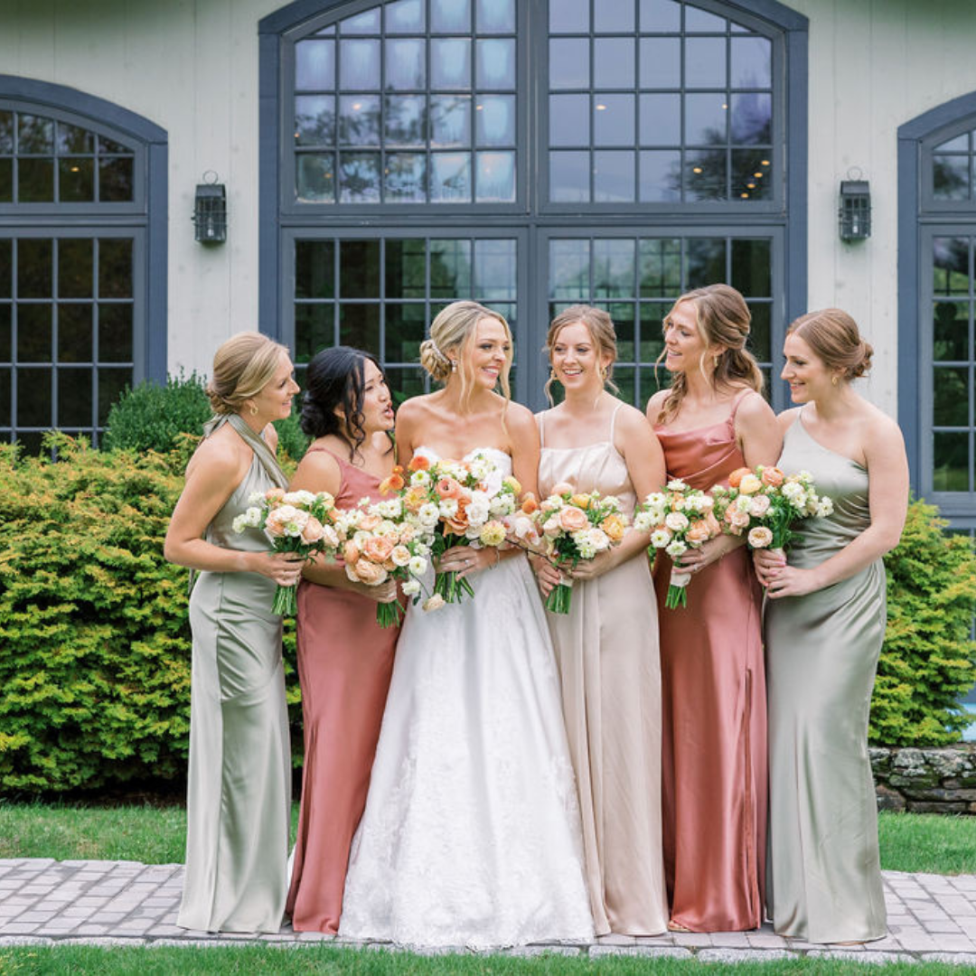 Bride and bridesmaids in elegant satin dresses holding floral bouquets, posing outside Winvian Farm in Connecticut before a luxury wedding.