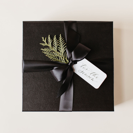 Luxury black holiday gift box with satin ribbon, evergreen sprig, and calligraphy gift tag reading “‘Tis the Season” – custom corporate holiday gift by Lavender + Pine
