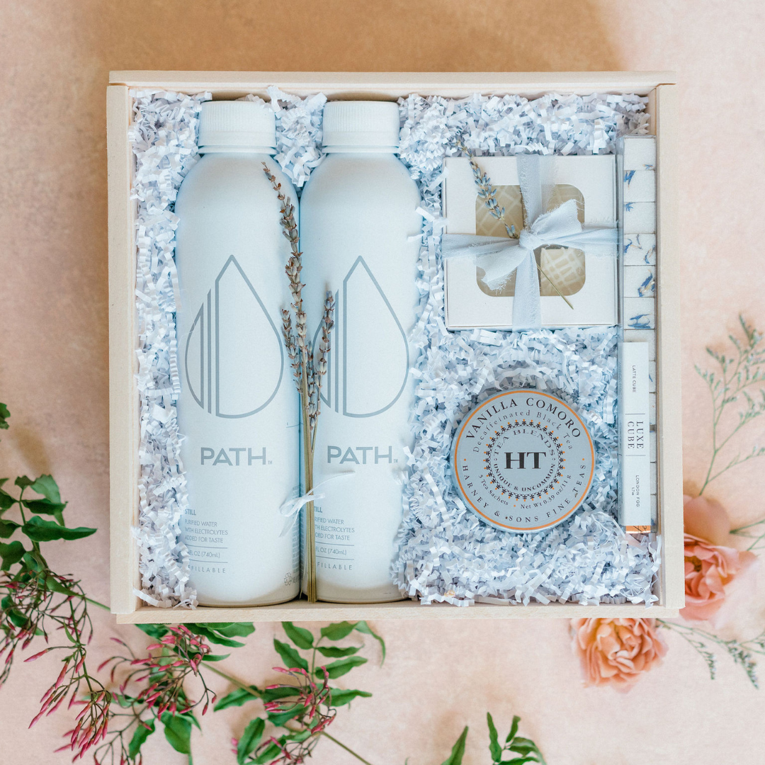 Curated wedding welcome gift box with PATH water bottles, Harney & Sons tea, and lavender accents, styled with pink florals by Lavender + Pine Gifting.