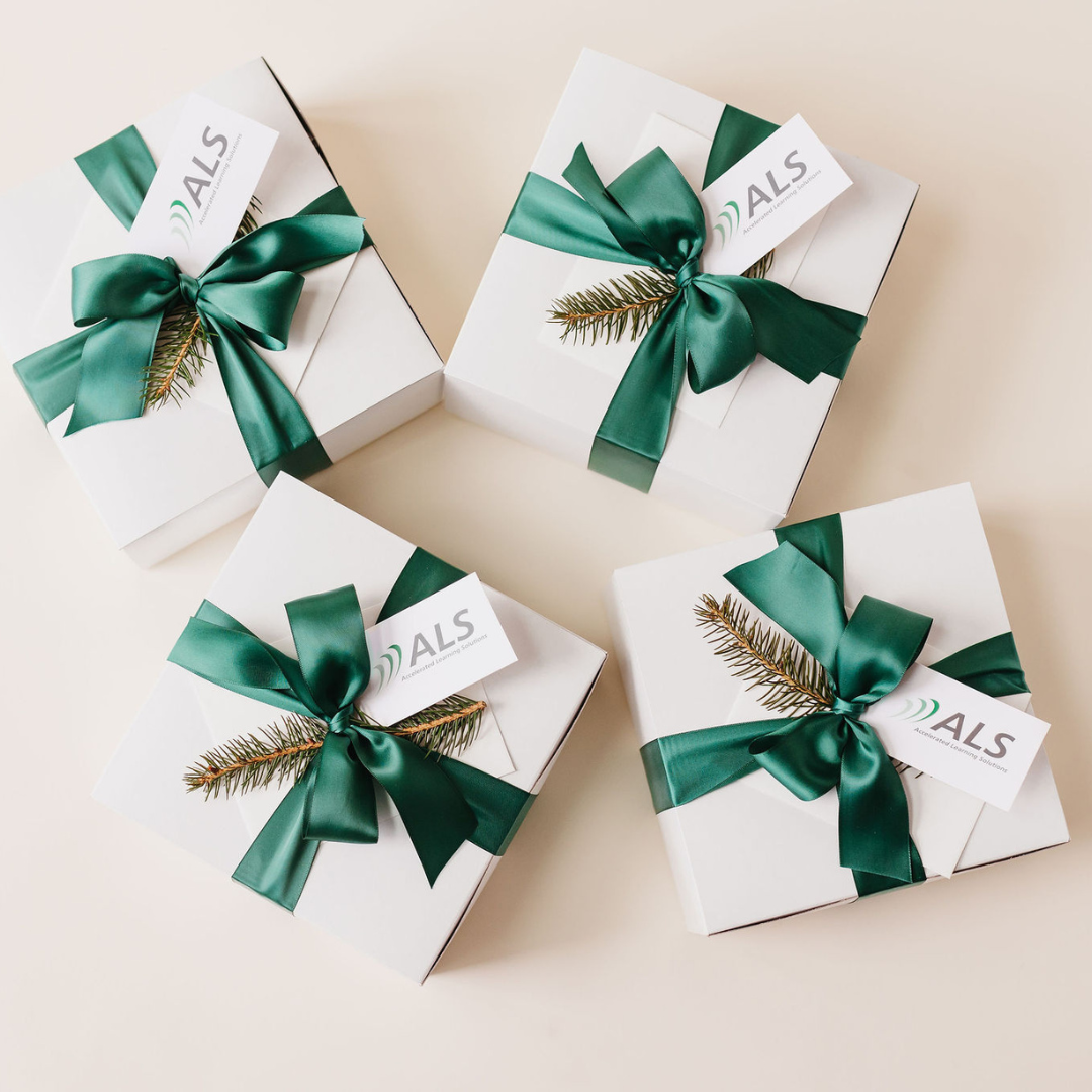 Flat lay of four white corporate gift boxes with dark green satin ribbons, each adorned with a branded ALS card and a pine sprig—styled for holiday gifting.