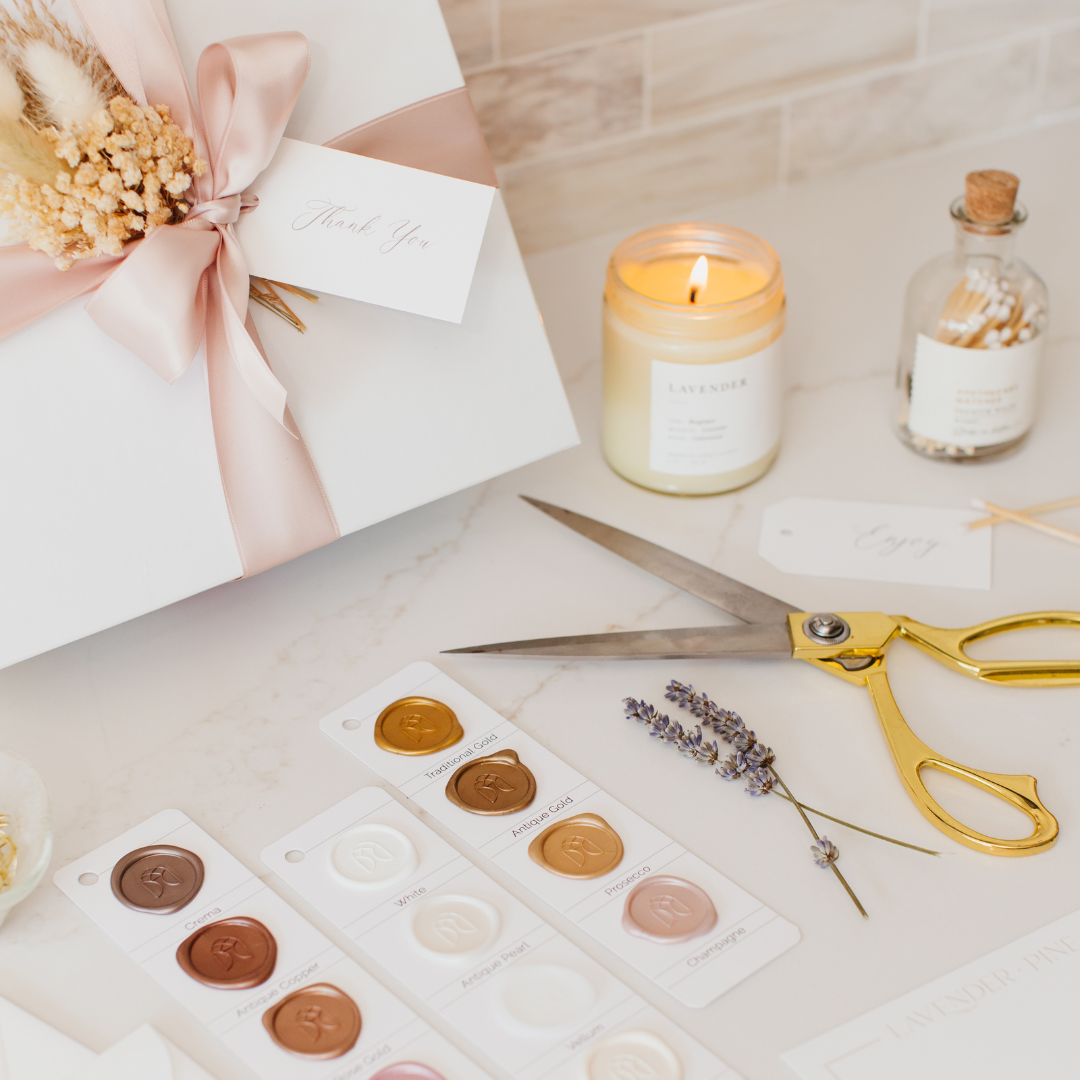 Styled flat lay of a white luxury gift box with blush satin ribbon, lavender candle, gold scissors, and wax seal swatches on a marble surface - Lavender + Pine Gifting