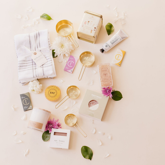 Flatlay of a curated Lavender + Pine gift box featuring gold measuring cups, tea, towel, bath salts, oat and honey soap, and wellness items, styled with florals and greenery.
