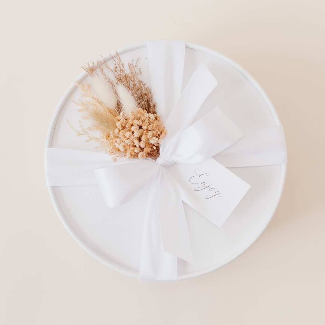 White round gift box tied with a white satin ribbon and dried floral arrangement, with a handwritten "Enjoy" gift tag - Lavender + Pine.