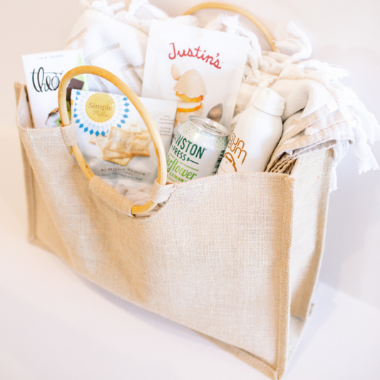 Neutral-toned wedding welcome gift bag filled with artisan snacks, a can of sparkling water, and a beach towel, curated by Lavender + Pine.