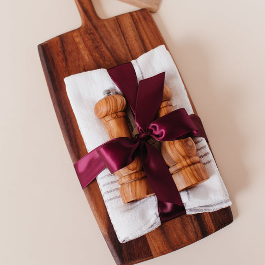 Closing gift with an acacia wood paddle cutting board, olive wood salt and pepper mills, and white striped tea towels wrapped in burgundy satin ribbon