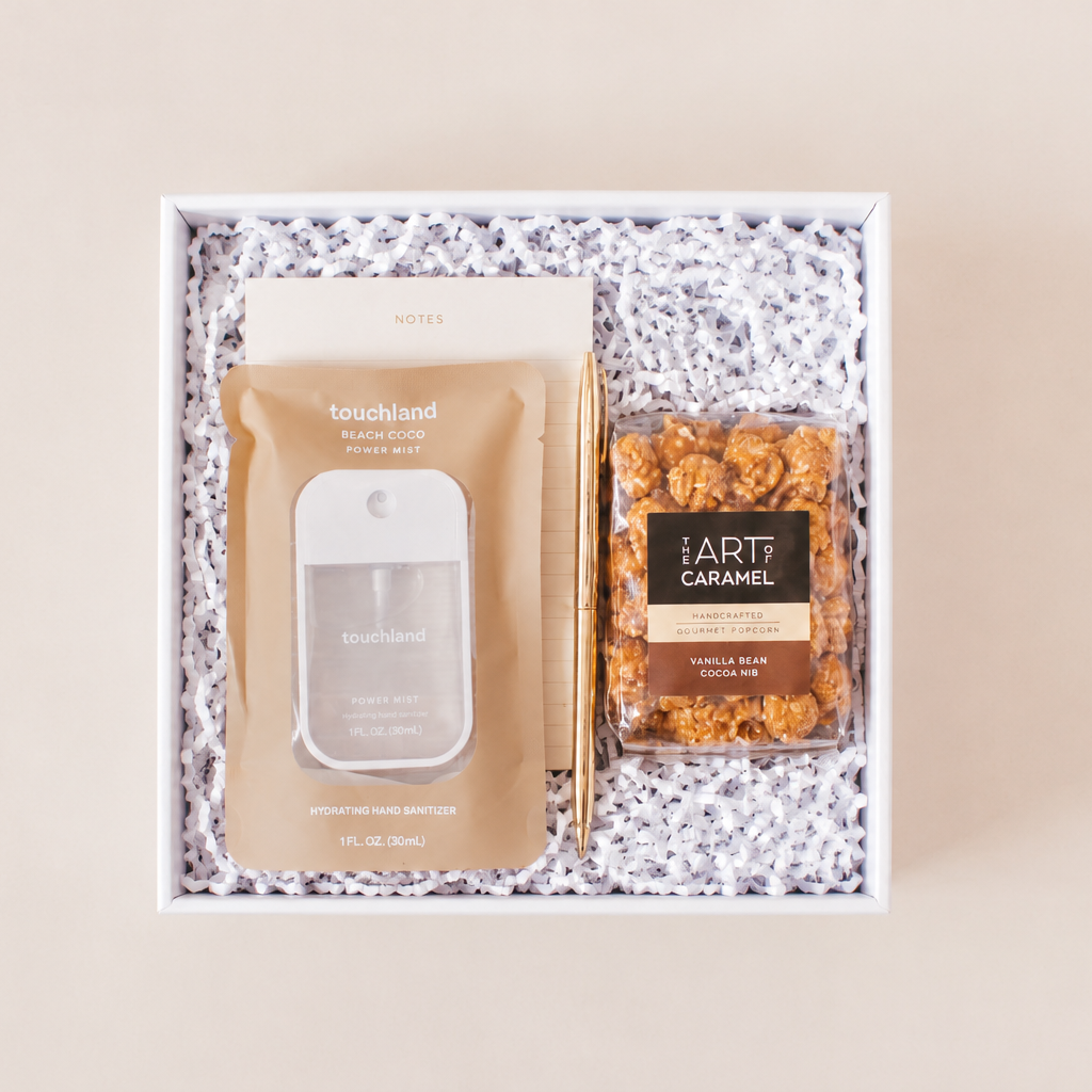 Desk essentials corporate gift box with Touchland Beach Coco hand sanitizer, notepad, gold pen, and The Art of Caramel vanilla bean cocoa nib gourmet caramel popcorn in a white box with crinkle paper