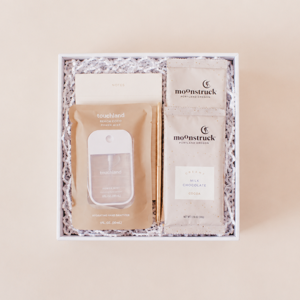 Desk essentials corporate gift box with Touchland Beach Coco hand sanitizer, notepad, gold pen, and two Moonstruck creamy milk chocolate hot cocoa packets in a white box with crinkle paper