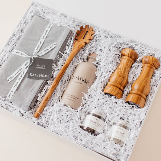 Lavender and Pine Entertainer's Kitchen Gift Box angle view featuring Kaf Home grey linen napkins, Le Marké olive oil, Natural OliveWood pasta spoon, Natural OliveWood salt and pepper mills, and La Boite grey salt and Tellicherry pepper in white magnetic gift box