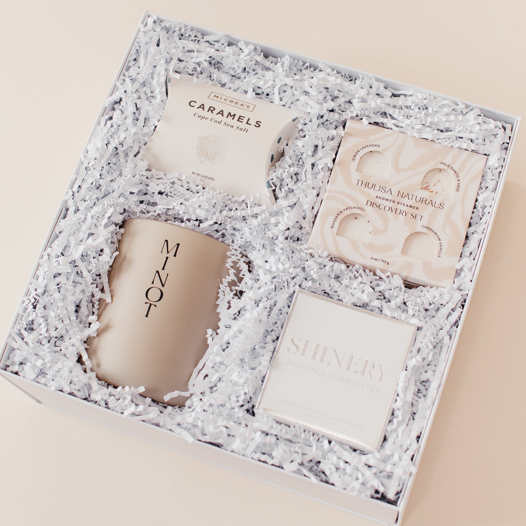 Lavender and Pine Refined Escape Gift Box angle view featuring McCrea's Cape Cod sea salt caramels, Thulisa Naturals shower steamer discovery set, Minot Cliffwalk candle, and Shinery radiance towelettes in white luxe two piece gift box