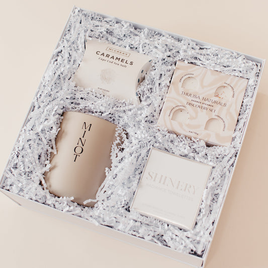 Lavender and Pine Refined Escape Gift Box angle view featuring McCrea's Cape Cod sea salt caramels, Thulisa Naturals shower steamer discovery set, Minot Cliffwalk candle, and Shinery radiance towelettes in white luxe two piece gift box