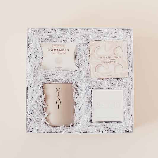 Lavender and Pine Refined Escape Gift Box featuring McCrea's Cape Cod sea salt caramels, Thulisa Naturals shower steamer discovery set, Minot Cliffwalk candle, and Shinery radiance towelettes in white luxe two piece gift box
