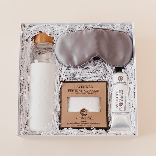 Lavender and Pine Soft Reset Gift Box featuring Soma glass water bottle, Pure Sol silk sleep mask, ElizabethW lavender exfoliating shower pouch, and ElizabethW lavender shea butter hand cream in white luxe two piece gift box
