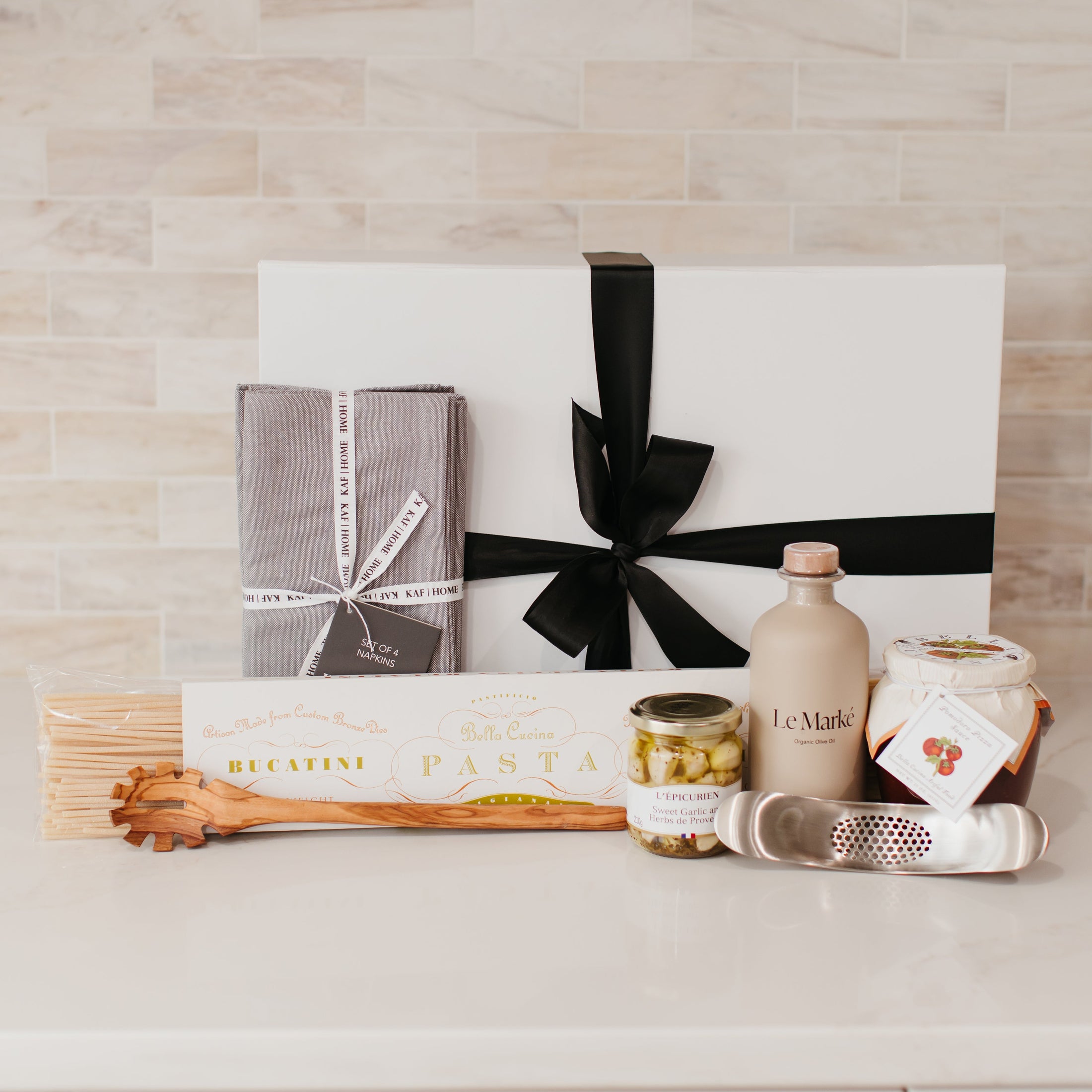 Lavender and Pine Sunday Sauce Gift Box detail view featuring Bella Cucina bucatini pasta, Le Marké olive oil, French Farm L'Epicurien sweet garlic and herbs, Bella Cucina arrabbiata sauce, stainless garlic rocker, Natural OliveWood pasta spoon, and Kaf Home grey linen napkins in white magnetic gift box