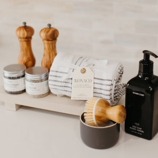 Lavender and Pine Well-Kept Kitchen Gift Box detail view featuring Natural OliveWood salt and pepper mills, La Boite grey salt and Tellicherry pepper, Monaco kitchen towels, Homecourt Cece dish soap, dish brush set, and Luxe B Co sandstone trivet riser