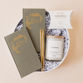 Load image into Gallery viewer, Lavender and Pine Written in Love Gift Box angle view featuring Seniman Calligraphy olive vow books, gold ballpoint pens, Roote lavender candle, and McCrea's Cape Cod sea salt caramels in white round gift box

